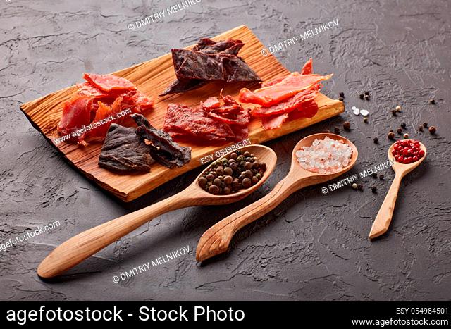 Jerky. Set of various kind of dried spiced meat on wooden tray, diverse peppercorns and salt on wooden spoons on dark gray background. Snack for beer