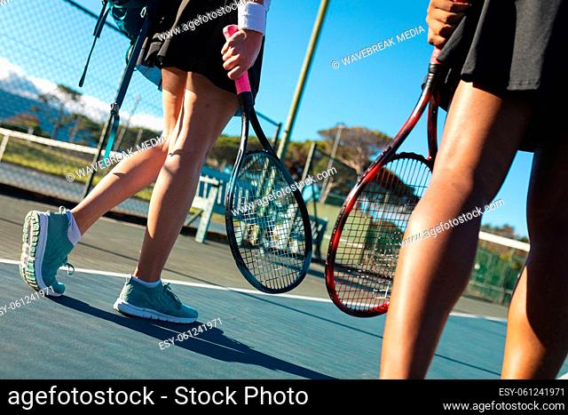 Low section of female multiracial tennis players walking rackets at court on sunny day