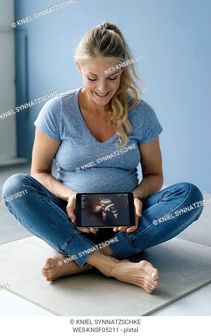 Smiling pregnant woman sitting on the floor showing ultrasound picture on tablet