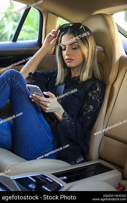 Young successful girl sitting in backseat of a luxury car on mobile phone while being driven around