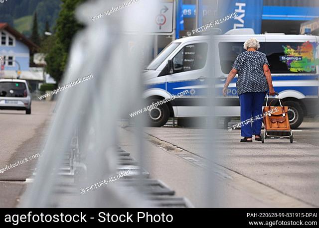 28 June 2022, Bavaria, Garmisch-Partenkirchen: An elderly woman walks with a shopping bag next to barriers and in front of a police car