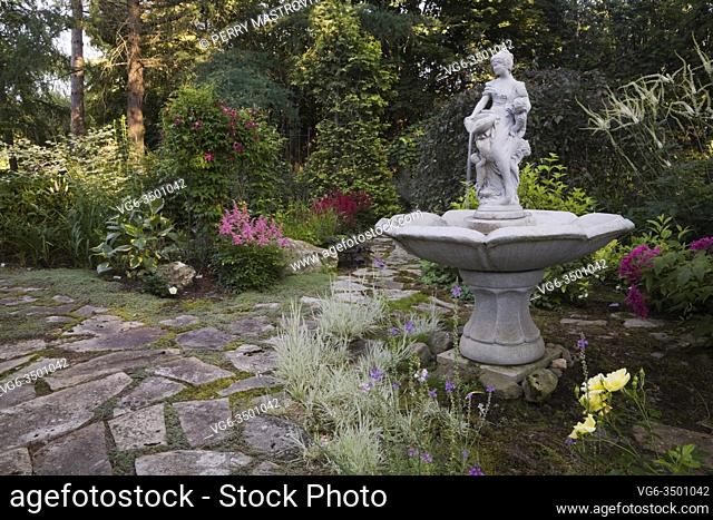 Flagstone path, water fountain and borders with ornamental grass plants, yellow Rosa â. . Flower Carpet' - Roses, Filipendula purpurea â