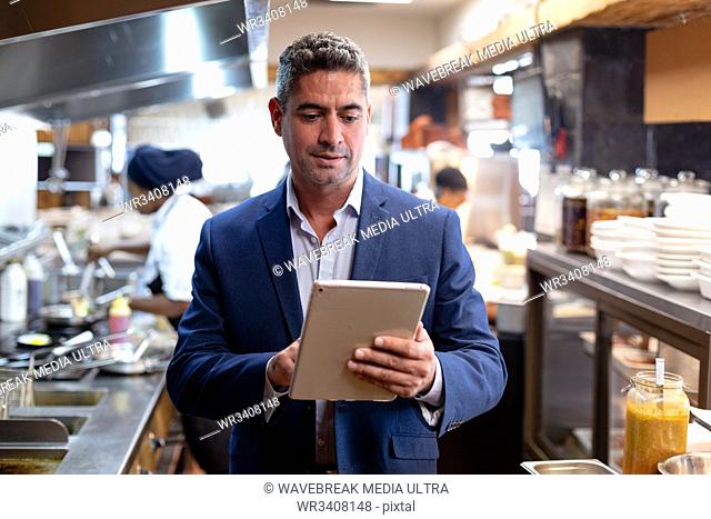 Front view close up of a middle aged Caucasian male restaurant manager using a tablet computer in a busy restaurant kitchen