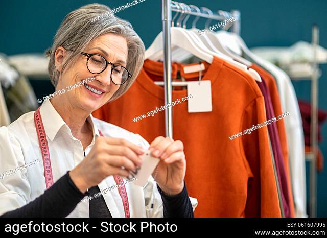 Tailor, work. Smiling gray-haired middle-aged woman in glasses hanging price tag on drawstring standing near rack with clothes in atelier
