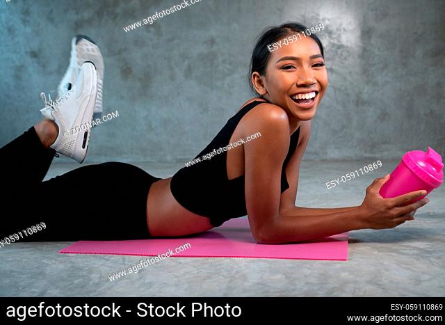 Happy healthy smiling woman holding protein shake relaxing on the pink yoga mat at the gym. Female in sportswear posing at fitness studio