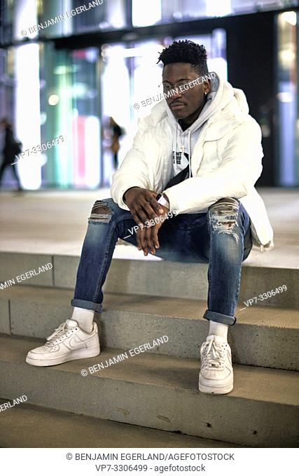 young stylish African man sitting on stairs at night in city, closed eyes, calm, resting, tired, in Munich, Germany
