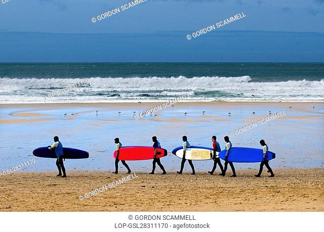 England, Cornwall, Newquay. Surfers walking in a line carrying their surfboards across Fistral Beach