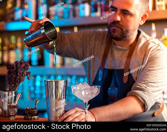 Young barman pouring fresh alcoholic drink from one metal glass into another on the bar counter