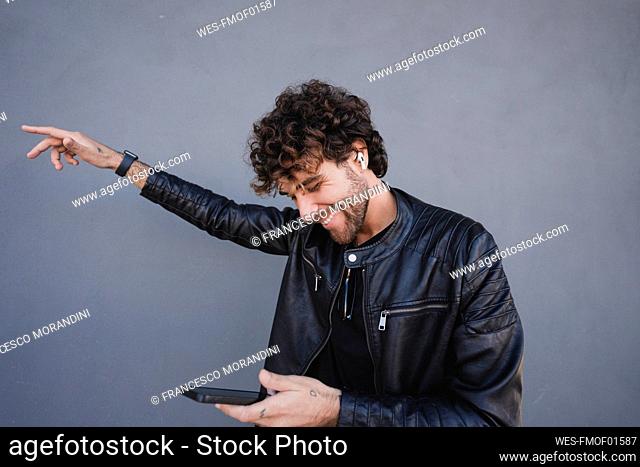 Happy man listening to music through wireless in-ear headphones holding smart phone dancing in front of gray wall