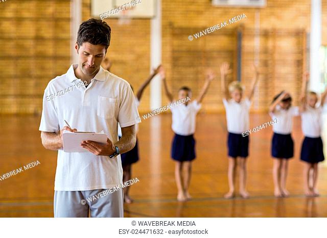 Sports teacher writing notes on clipboard while students exercising