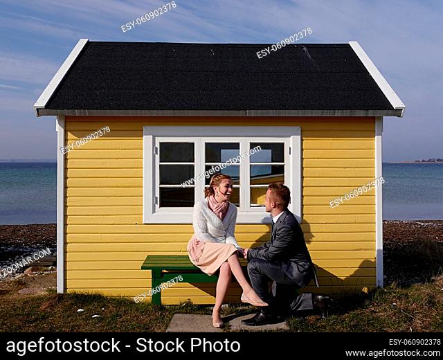 An attractive happy young man on one knee proposing to his beautiful girlfriend in front of cute, yellow beach hut for fun on Danish island of Aero