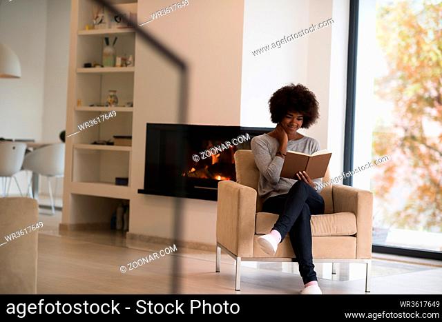 Young beautiful african american woman sitting in front of fireplace at home on a cold autumn day and reading book