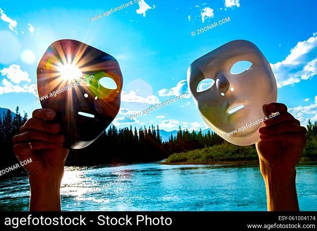 Light and dark concept in nature as a person holds two masks against the sun over a large lake by mountains, with lens flare as rays shine through eye