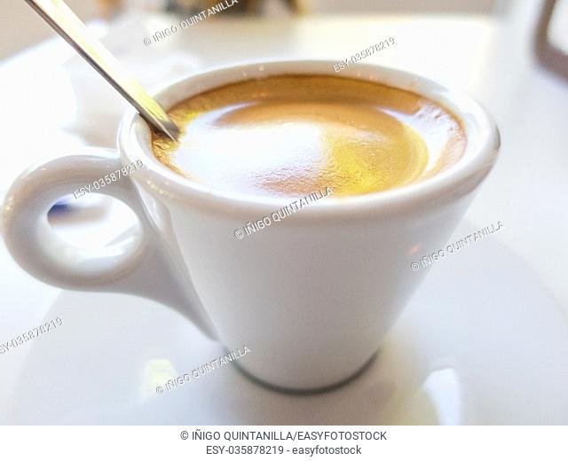 macro of white small cup with espresso coffee, with metal spoon, on table of restaurant bar or cafe