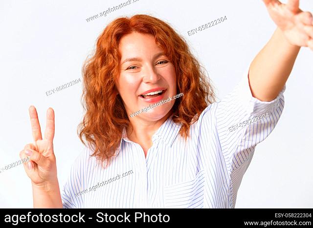 People, emotions and lifestyle concept. Close-up of smiling attractive middle-aged woman with red curly hair showing peace sign and taking selfie