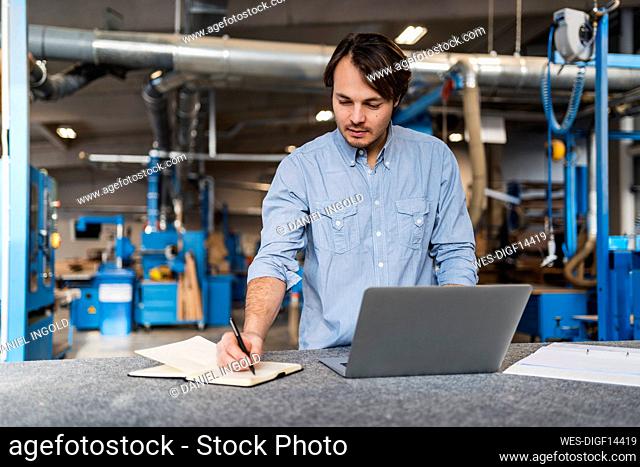 Male expertise with laptop writing in book while standing at industry