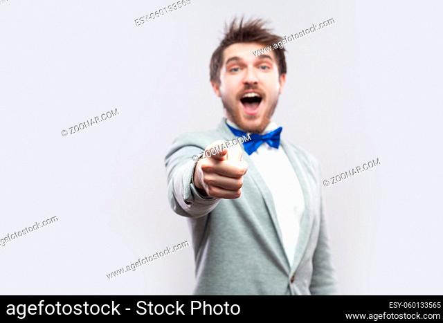Portrait of surprised handsome bearded man in casual grey suit and blue bow tie standing pointing and looking at camera with amazed face