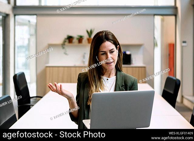 Businesswoman with in-ear headphones gesturing during video call on laptop at office