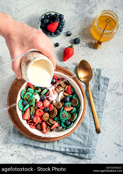 Trendy food - pancake cereal. Milk pouring on heap of multicolor mini cereal pancakes in bowl. Tiny cereal pancakes with berries in craft plate over gray cement...