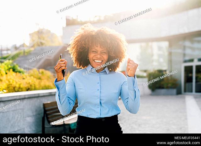 Happy Afro businesswoman holding mobile phone celebrating on sunny day