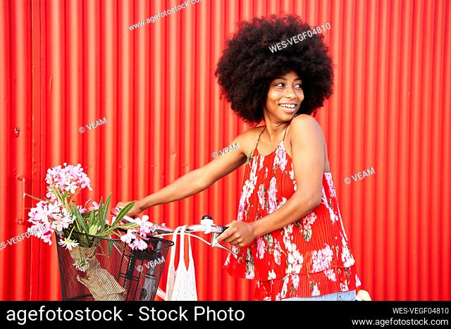 Afro woman with bicycle smiling while standing by red wall