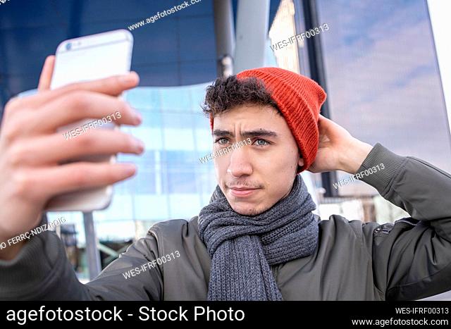 Young man adjusting knit while taking selfie through smart phone in city