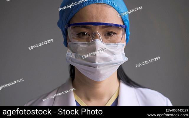 Doctor in safety glasses and mask. Portrait of asian woman in medical uniform and goggles looking at camera on gray background