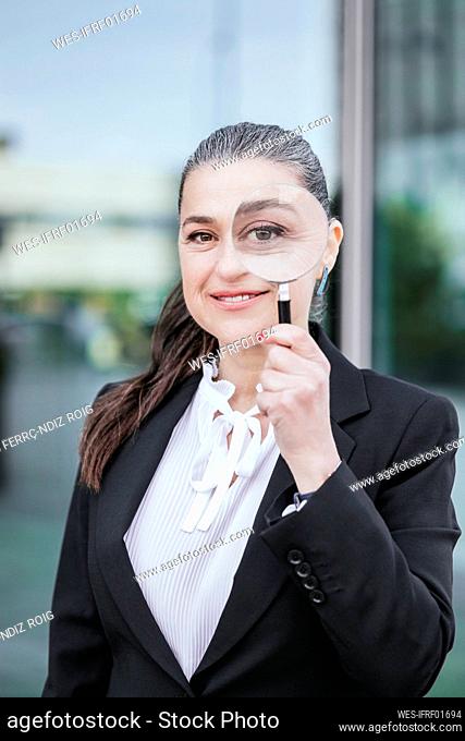 Smiling businesswoman looking through magnifying glass in front of glass wall