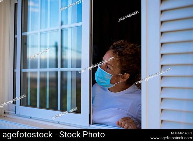 portrait of woman looking outdoor from the window of her house in lockdown or quarantine to be safe from the coronavirus or covid-19 - fase 1 or 2 of lockdown