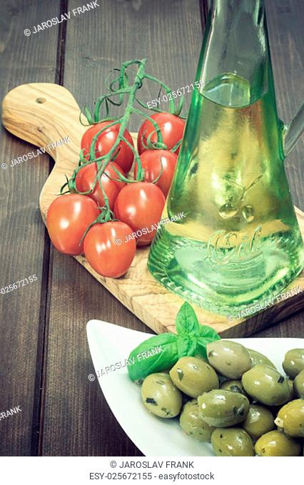 White bowl with marinated green olives is placed on a wooden desk. Tomatoes and bottle of oil are lying on a desk of olive wood