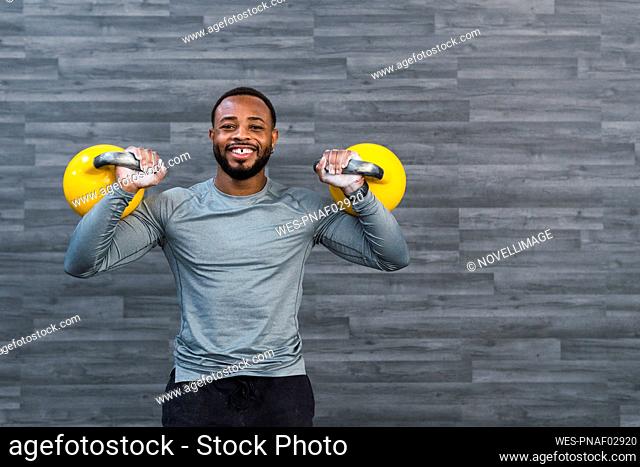 Smiling sportsman carrying kettlebells in front of wall at gym
