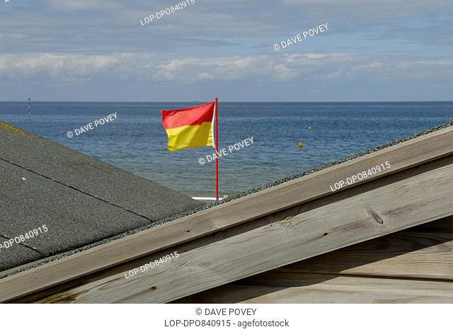 England, Kent, Minnis Bay, A bathing flag seen between the roofs of two beach huts. The bathing flag marks one end of a section of beach patrolled by lifeguards