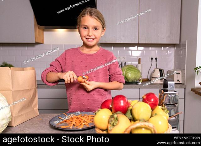Portrait of smiling girl peeling carrots in kitchen at home