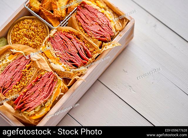 Enormous sandwiches with pastrami beef in wooden box. Served with baked potatoes, pickles and french mustard. Top view, flat lay