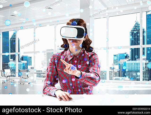 Conceptual image of young woman in checkered shirt using virtual reality headset with media interface while sitting inside bright building