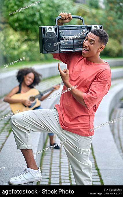 Meloman. Cheerful dark-skinned guy standing pointing at record-player holding on his shoulder and girl sitting behind with guitar outdoor