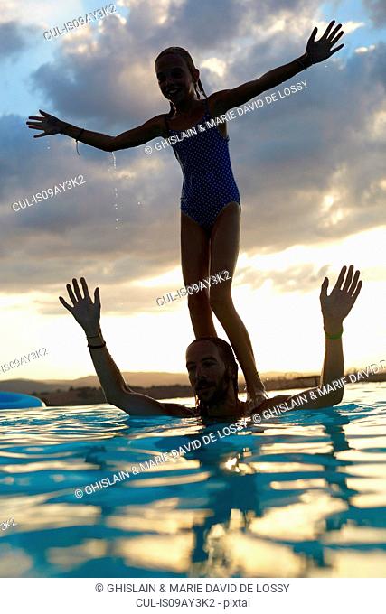 Silhouetted girl balancing on fathers shoulders in swimming pool, Buonconvento, Tuscany, Italy