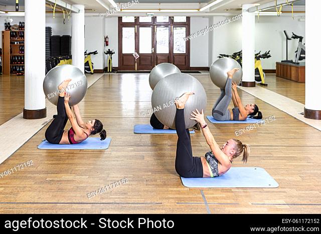 Side view of barefoot female athletes in activewear lying on floor and raising fit balls in legs and arms during fitness workout in daytime in gym
