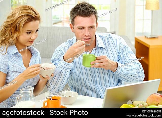 Couple having healthy breakfast at home, eating cereals drinking coffee, and looking at laptop computer