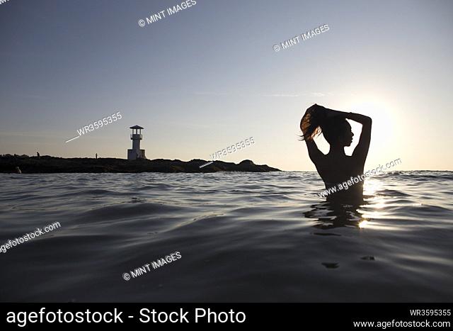 Rear view of woman bathing in the ocean at sunset, lighthouse in the distance