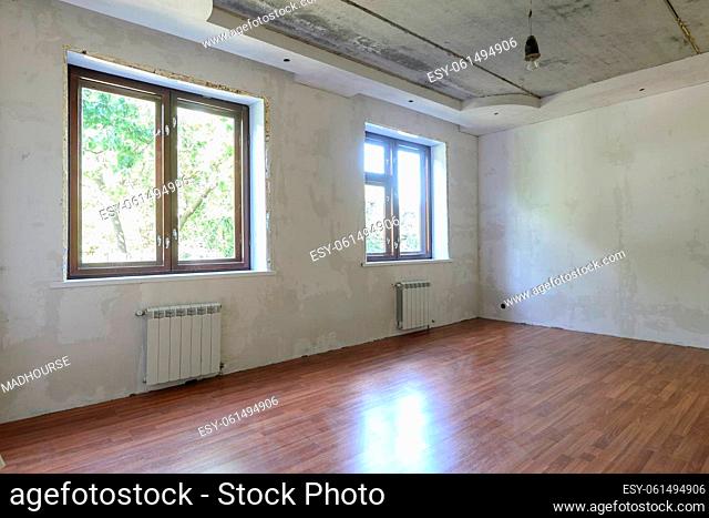 The interior of an empty room during renovation, there are two large windows in the room, heating radiators are located under the windows