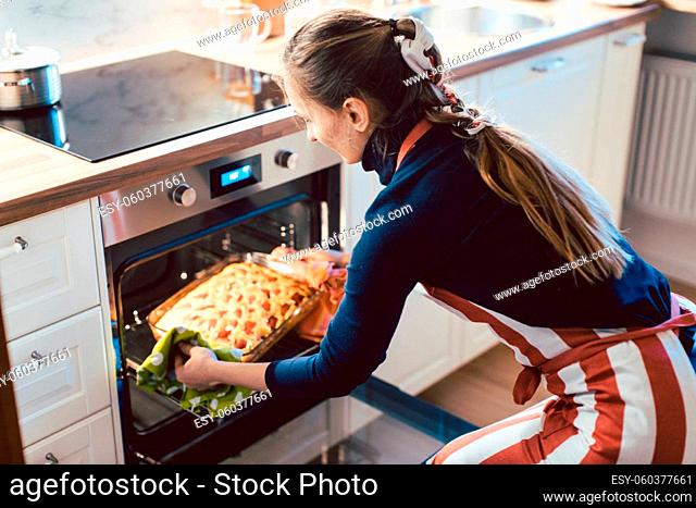 Beautiful young woman wearing apron removing baked apple pie from microwave oven in kitchen at home