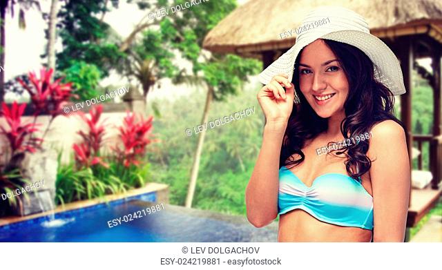 people, fashion, summer holidays, travel and tourism concept - happy young woman in bikini swimsuit and sun hat over hotel resort background