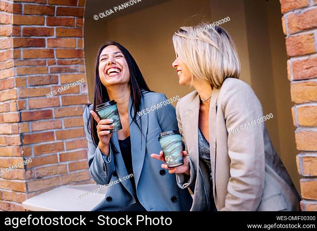 Cheerful female friends enjoying coffee sitting together at wall