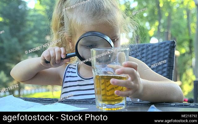 Cute little girl carefully looks into the lens at the juice in the glass. Close-up of blonde girl studying her juice in glass while looking at her through...