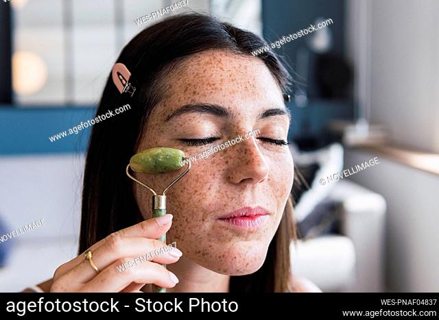 Young woman with freckle face massaging using jade stone roller at home
