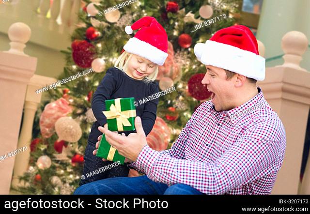 Happy young girl and father wearing santa hats opening gift box in front of decorated christmas tree