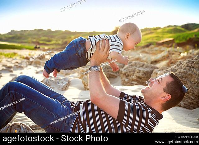 Young father playing with baby son as lying at the beach, smiling happy having fun. Side view