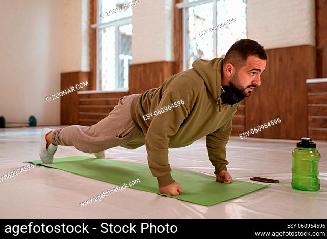 Young handsome sports guy doing push ups exercise in empty gym with green bottle of water next to him all in khaki color clothes