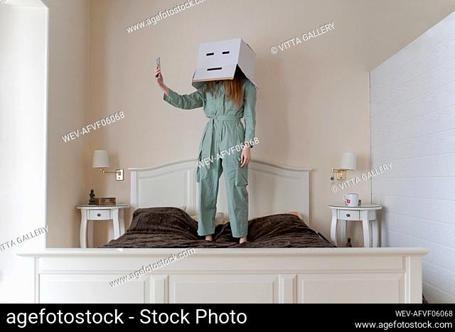 Woman wearing a cardbox on head with bored smiley taking a selfie on her bed